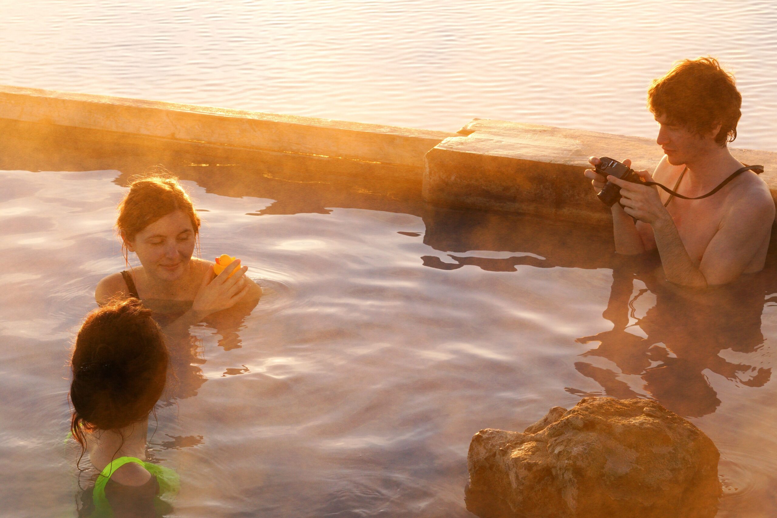Group of people in a rocky pool during sunset; one person on the right holding a camera.