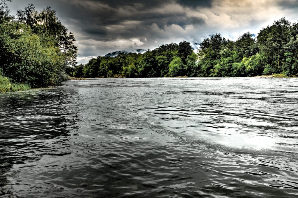 Wide river with rippled water flanked by dense green trees under dark stormy clouds overhead.