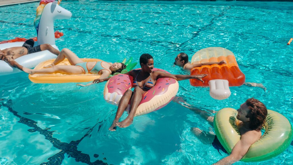 Group of friends relaxing on colorful inflatable pool toys in a bright blue swimming pool.