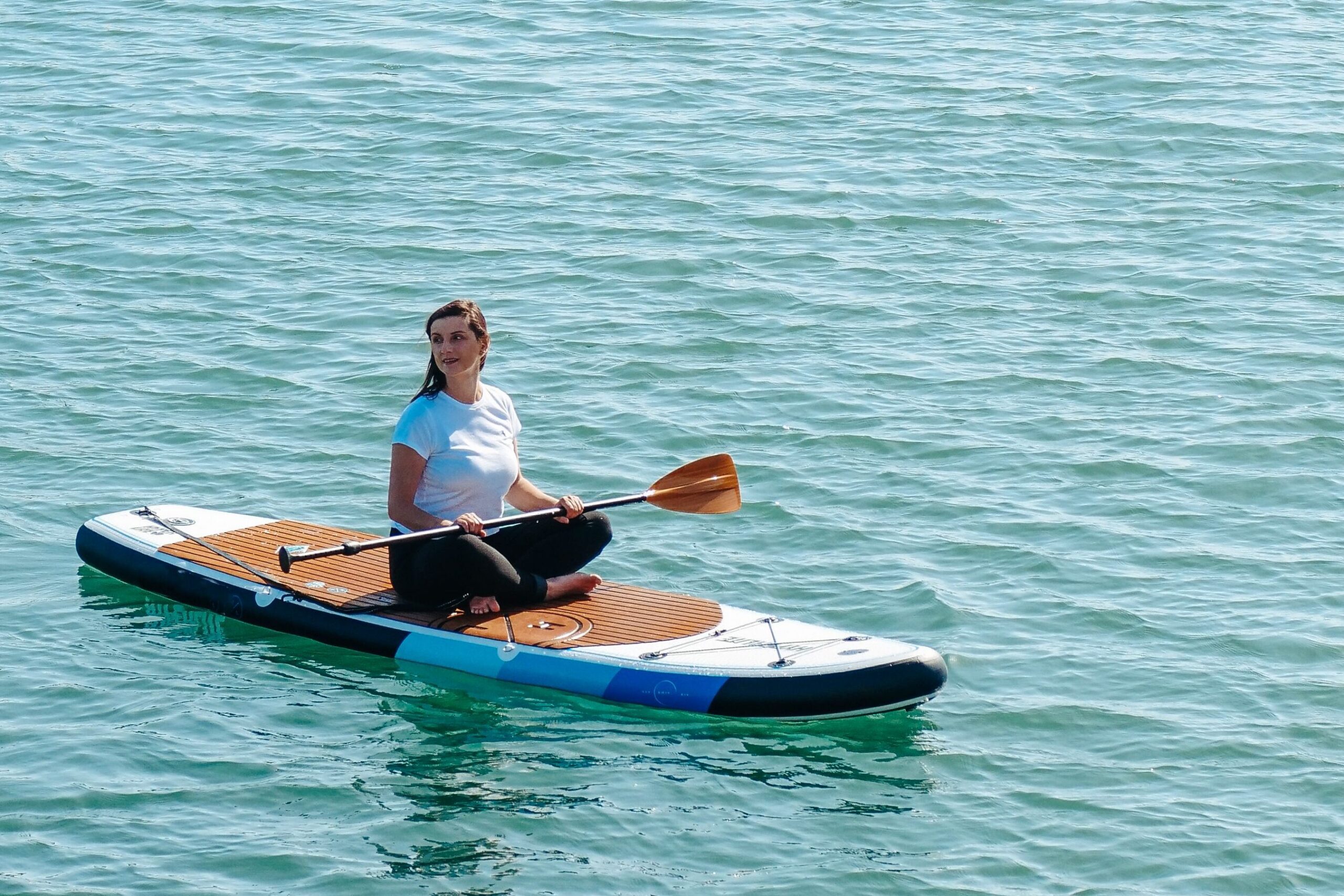 Woman paddling on a brown and blue stand-up paddleboard in calm turquoise water near a rocky breakwater in the distance at the horizon.